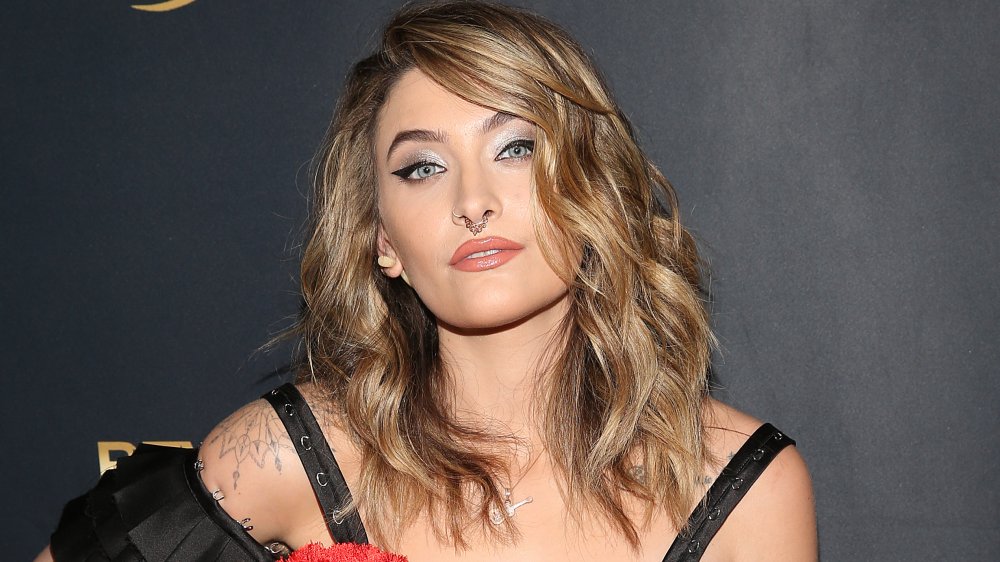 Paris Jackson con cabello más largo, anillo en la nariz y vestido rojo y negro, haciendo una pose en la alfombra roja