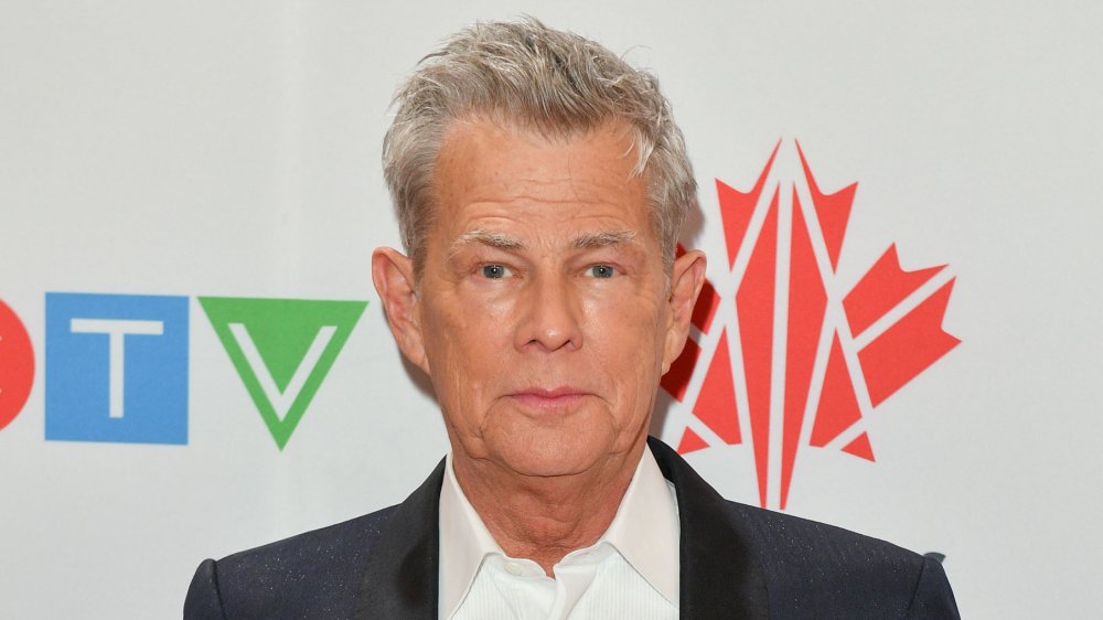 David Foster posando con expresión neutral en una alfombra roja, vistiendo un blazer negro y una camisa de vestir blanca