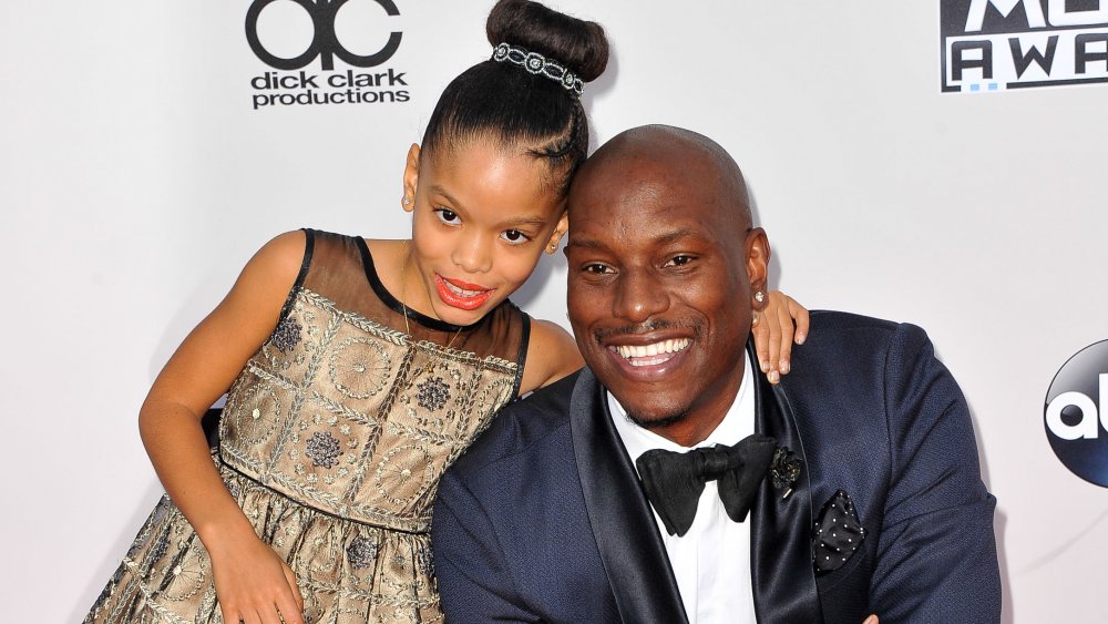 Tyrese Gibson con un traje azul, sonriendo y arrodillado en la alfombra roja junto a su hija Shayla Gibson