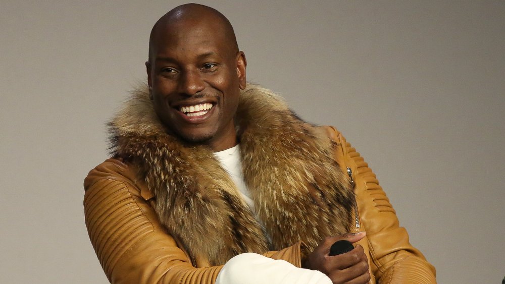 Tyrese Gibson sonriendo grande, vistiendo una chaqueta de cuero marrón claro durante una entrevista