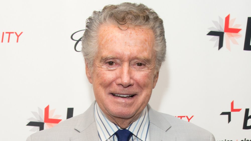 Regis Philbin en traje marrón y corbata azul, con una pequeña sonrisa en un evento de alfombra roja