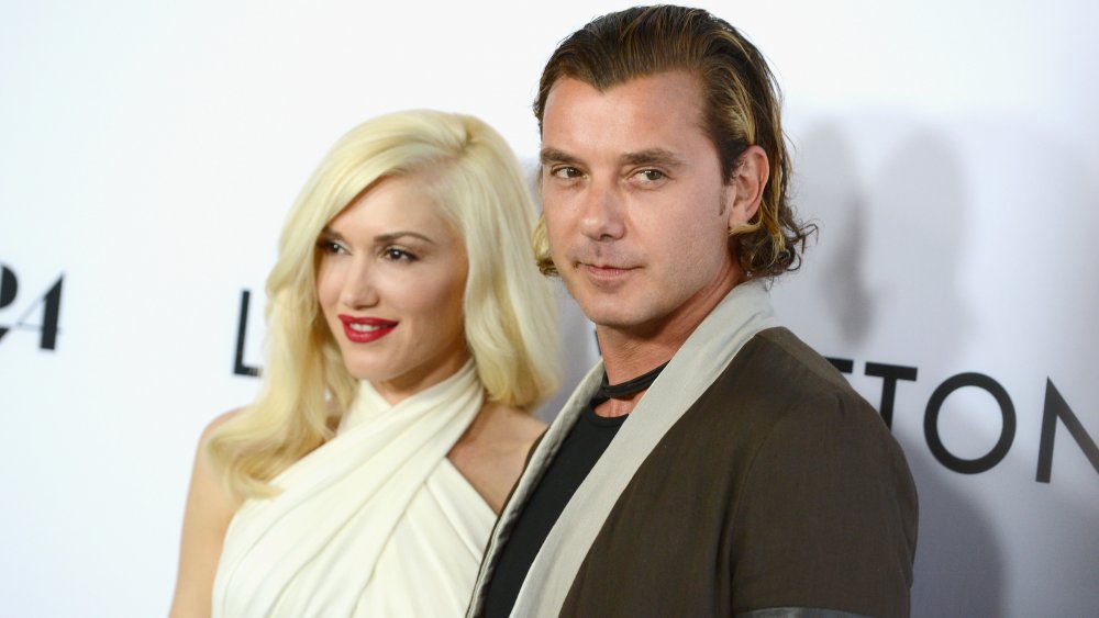 Gwen Stefani sonriendo con un vestido blanco con top corto y Gavin Rossdale con una expresión seria en un blazer marrón