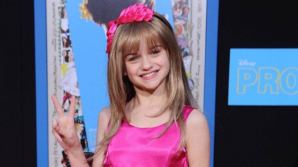 Joey King dando un signo de paz y sonriendo en la alfombra roja, con un vestido rosa y una diadema