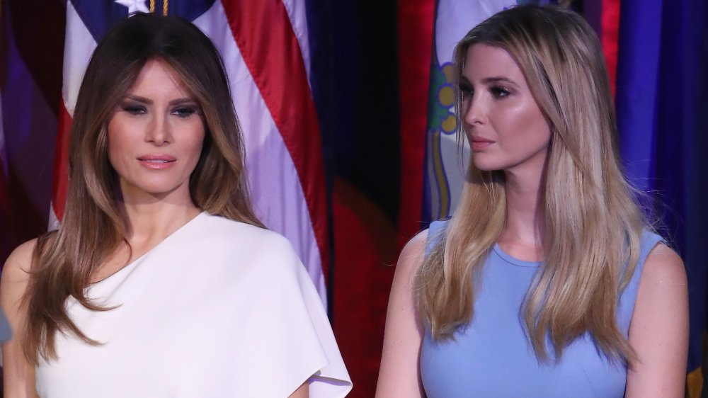 Melania Trump con un vestido blanco, Ivanka Trump con un vestido azul, de pie uno al lado del otro con expresiones serias