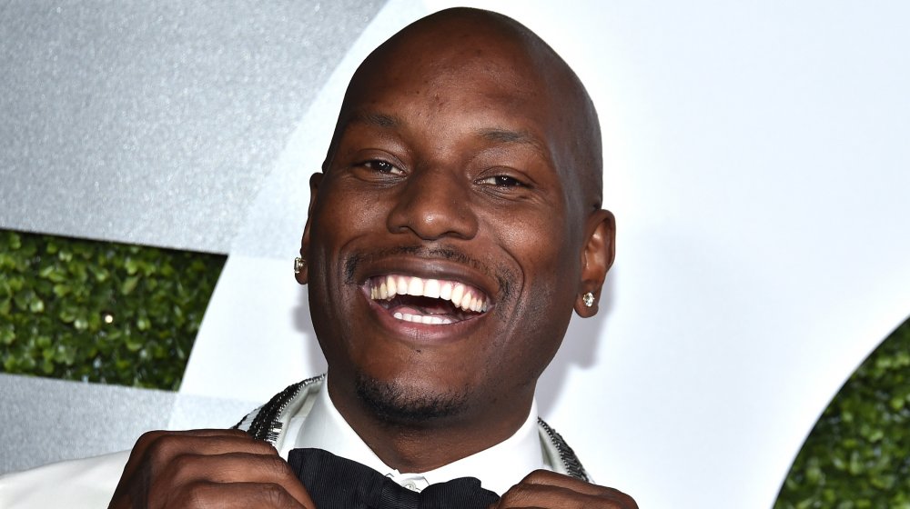 Tyrese Gibson con un traje blanco, sonriendo ampliamente mientras ajusta su pajarita negra en la alfombra roja