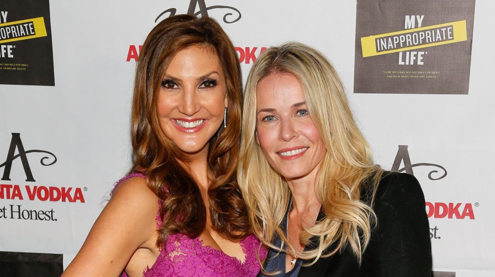 Heather McDonald con un vestido morado de encaje y Chelsea Handler con un traje negro, sonriendo en un evento de fiesta del libro en 2013