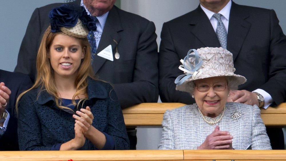 La princesa Beatriz con un traje azul oscuro y un sombrero, la reina Isabel con un traje azul claro, aplaudiendo y sonriendo