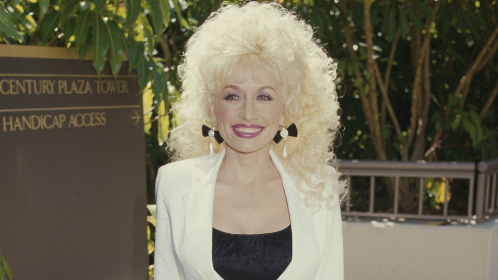 Dolly Parton sonriendo a lo grande con un traje blanco y una blusa negra en 1985 a los 39 años