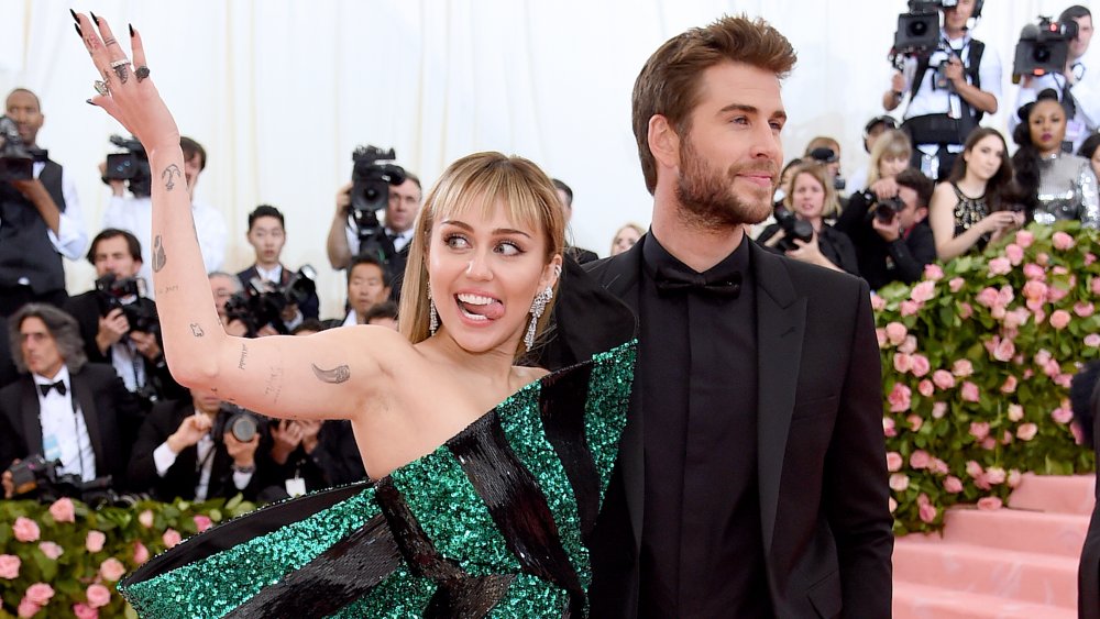 Miley Cyrus con un vestido verde y negro, sonriendo con la lengua fuera mientras Liam Hemsworth, con un traje completamente negro, mira hacia otro lado.