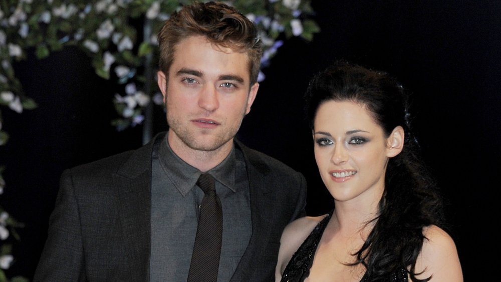 Robert Pattinson con una expresión neutra con un traje gris oscuro, Kristen Stewart sonriendo con un vestido negro