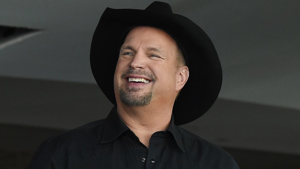 Garth Brooks con una camisa negra abotonada y sombrero de vaquero, sonriendo mientras mira hacia un lado