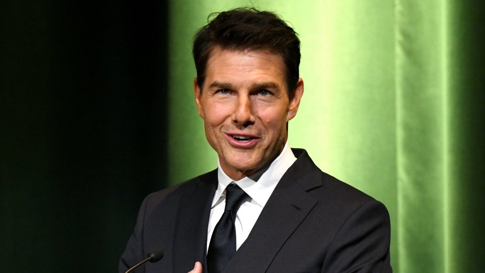 Tom Cruise hablando en un podio, frente a la pared verde