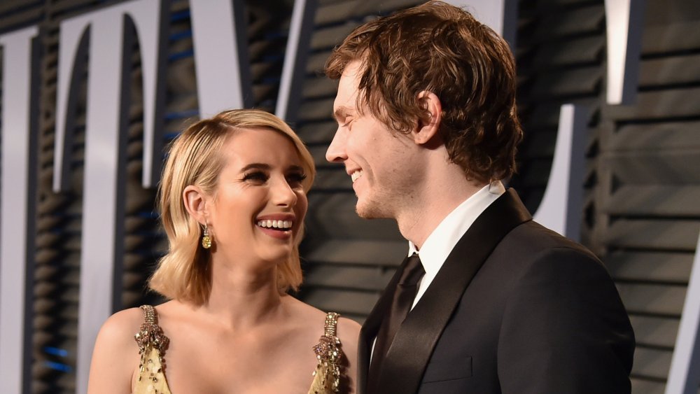 Emma Roberts con un vestido dorado y Evan Peters con un traje negro, sonriendo el uno al otro en la alfombra roja