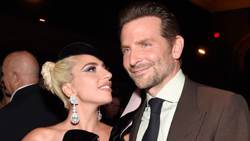 Lady Gaga con un vestido negro y un sombrero, sonriendo y mirando a un sonriente Bradley Cooper