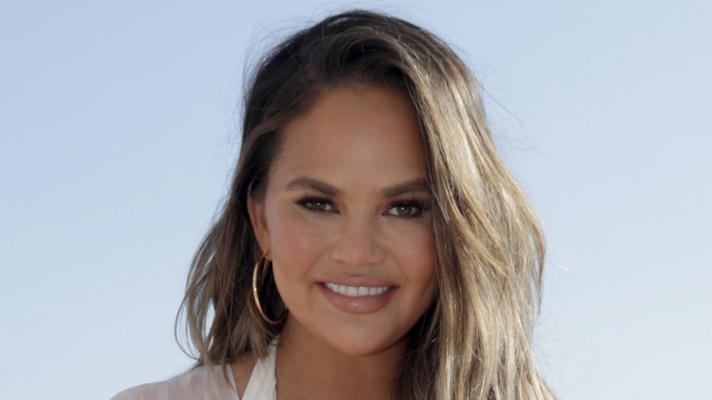 Chrissy Teigen con un vestido blanco y pendientes de aro de oro, sonriendo en la playa