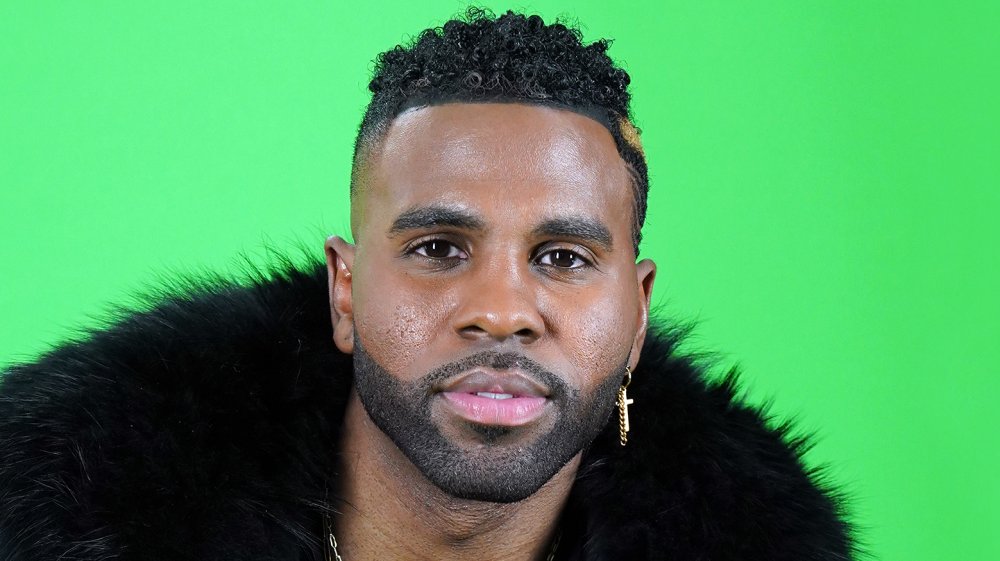 Jason Derulo con un abrigo de piel negro y un pendiente de cruz de oro, frente a una pantalla verde