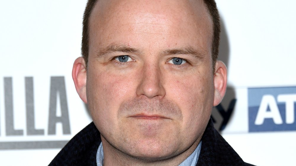 Rory Kinnear con un abrigo a cuadros negro y azul, posando con una expresión neutra