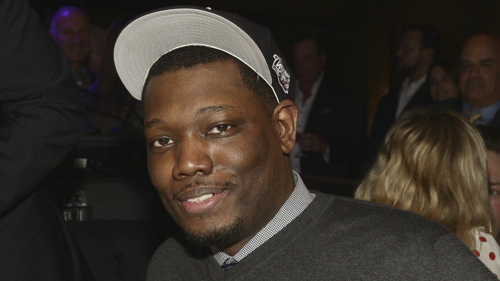 Michael Che con un suéter gris y sombrero negro, posando con una pequeña sonrisa