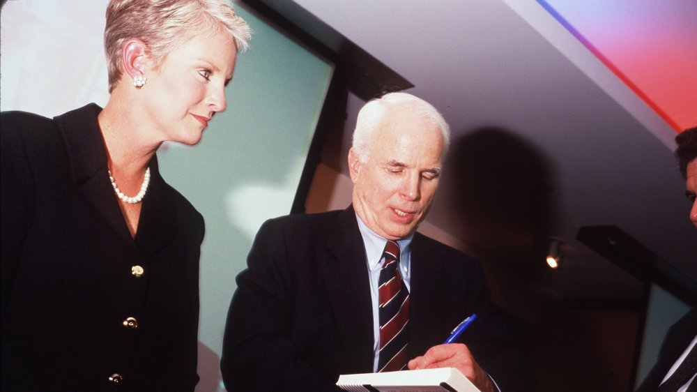 Cindy McCain con un vestido de blazer negro y perlas, viendo como su marido John McCain firma un libro en 1999