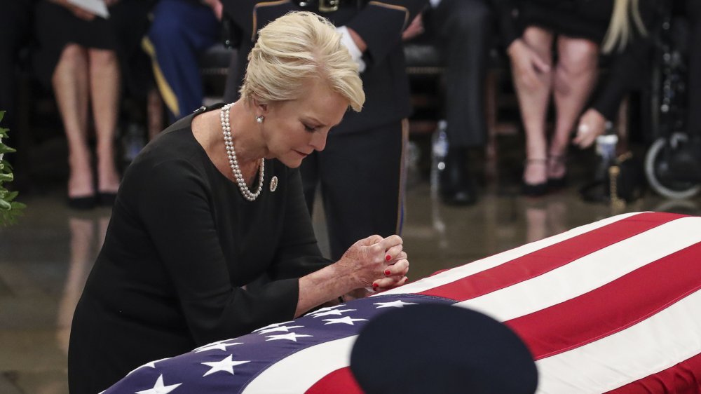 Cindy McCain vistiendo de negro y perlas, arrodillada y rezando sobre el ataúd de John McCain