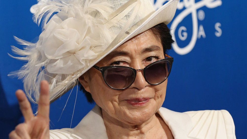 Yoko Ono con un blazer blanco, sombrero y gafas de sol, sonriendo y dando el signo de paz