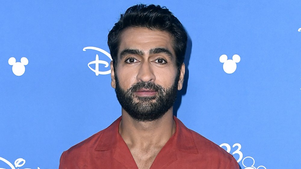 Kumail Nanjiani en un botón rojo, posando en un evento de Disney con una expresión neutral