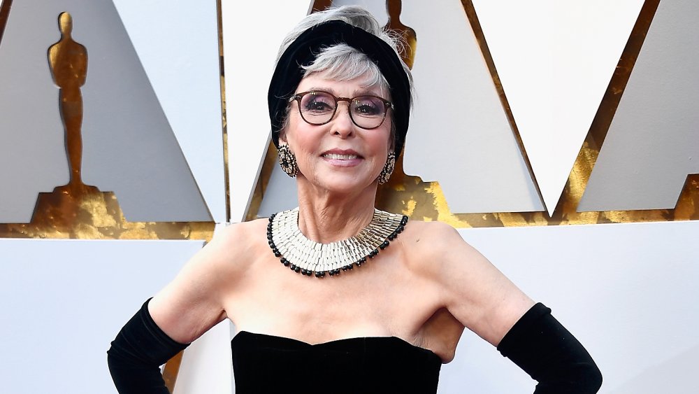 Rita Moreno con un vestido negro, sonriendo y posando con ambas manos en sus caderas en los Oscar