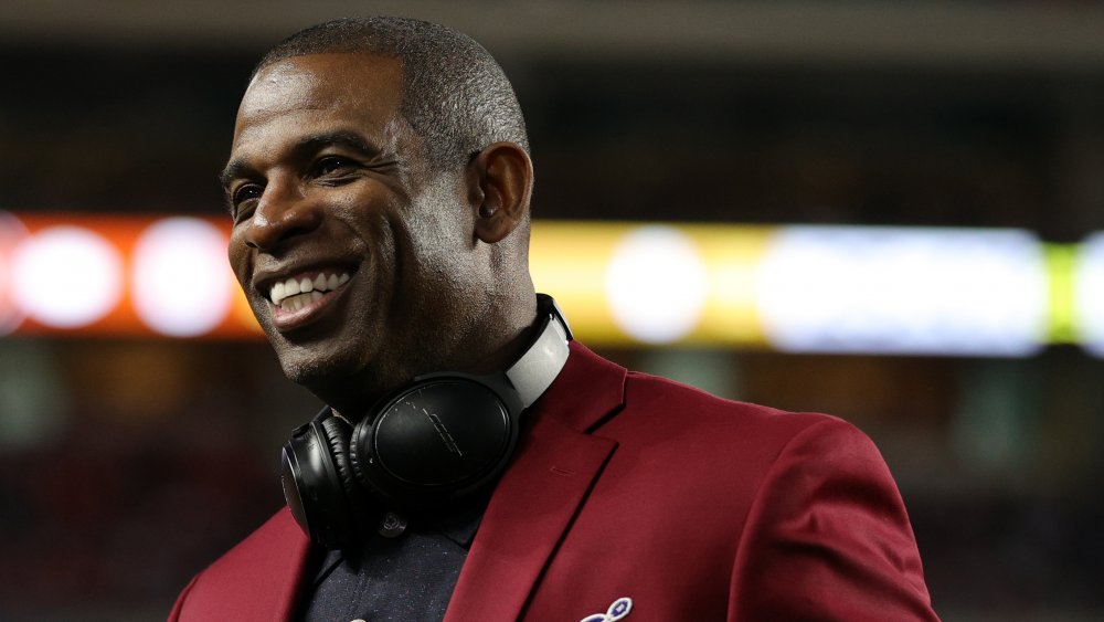 Deion Sanders sonriendo, mientras mira hacia un lado, en un blazer granate y polo negro