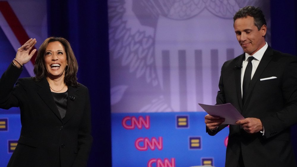 Kamala Harris saludando en el escenario y Chris Cuomo mirando sus notas junto a su