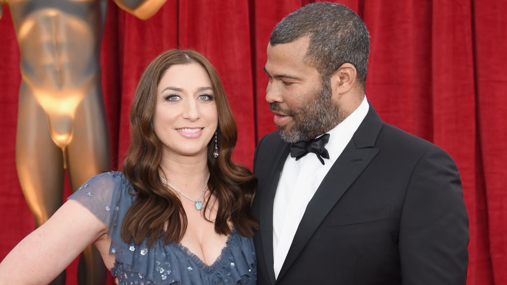 Chelsea Peretti sonriendo con un vestido azul, Jordan Peele en un traje negro mirando a su