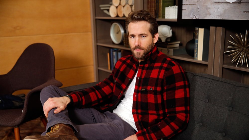 Un Ryan Reynolds barbudo sentado con la pierna cruzada, vestido a cuadros 