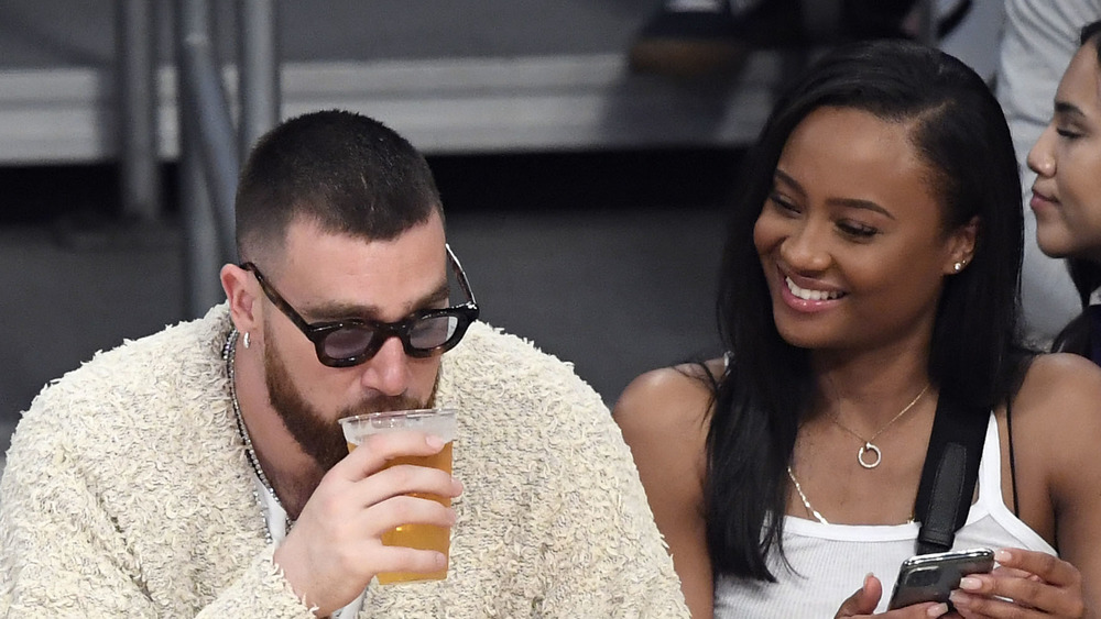 Travis Kelce, bebiendo una cerveza, en la cancha en un partido de la NBA, con gafas de sol;  Kayla Nicole, cabello largo suelto, sonriendo a Travis Kelce, camiseta blanca sin mangas, lado de la cancha en un juego de baloncesto