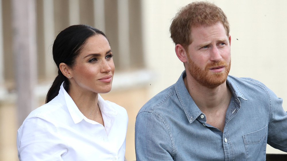 Meghan Markle vestida de blanco se sienta junto al príncipe Harry con una camisa de mezclilla mientras miran pensativamente un evento