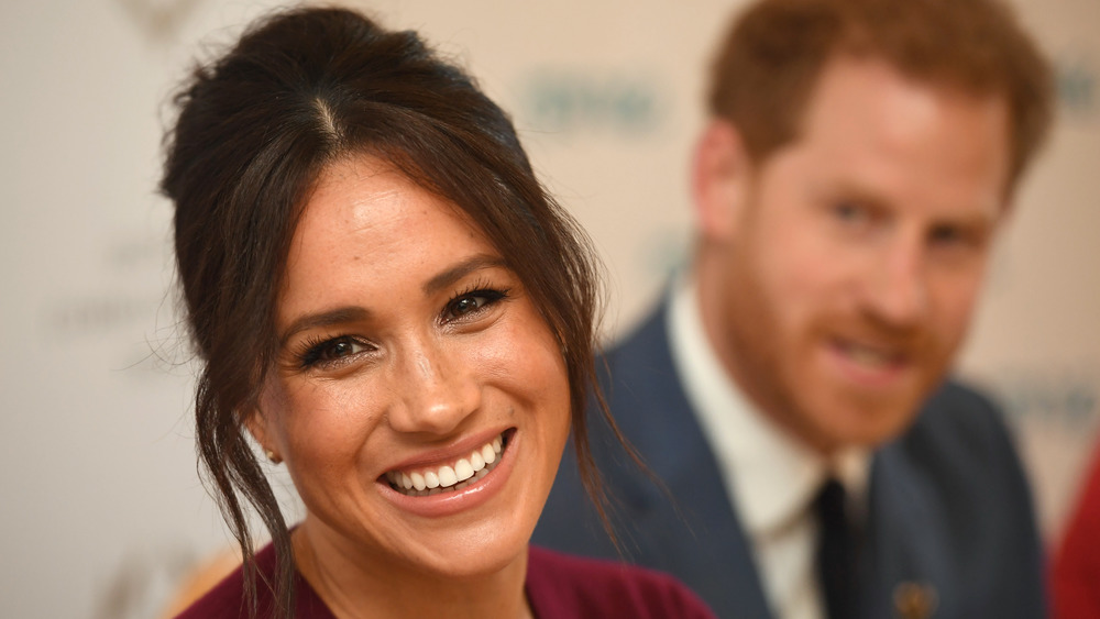 Meghan Markle sonriendo con el príncipe Harry de fondo