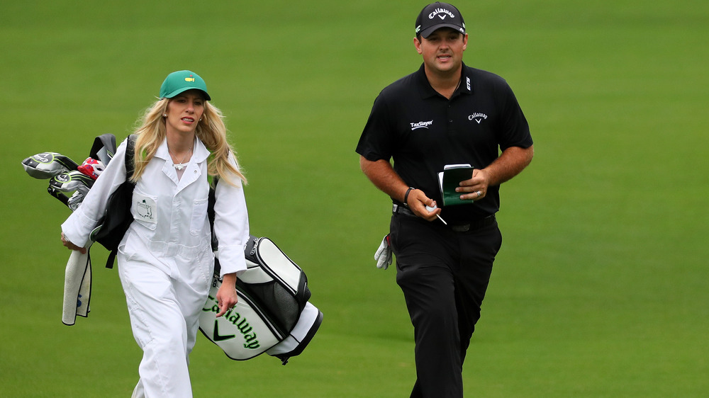 Justine Reed, trabajando como caddie, vestida de blanco, vestida con un odio, sosteniendo palos de golf;  Patrick Reed, vestido todo de negro y sombrero, en el campo de golf