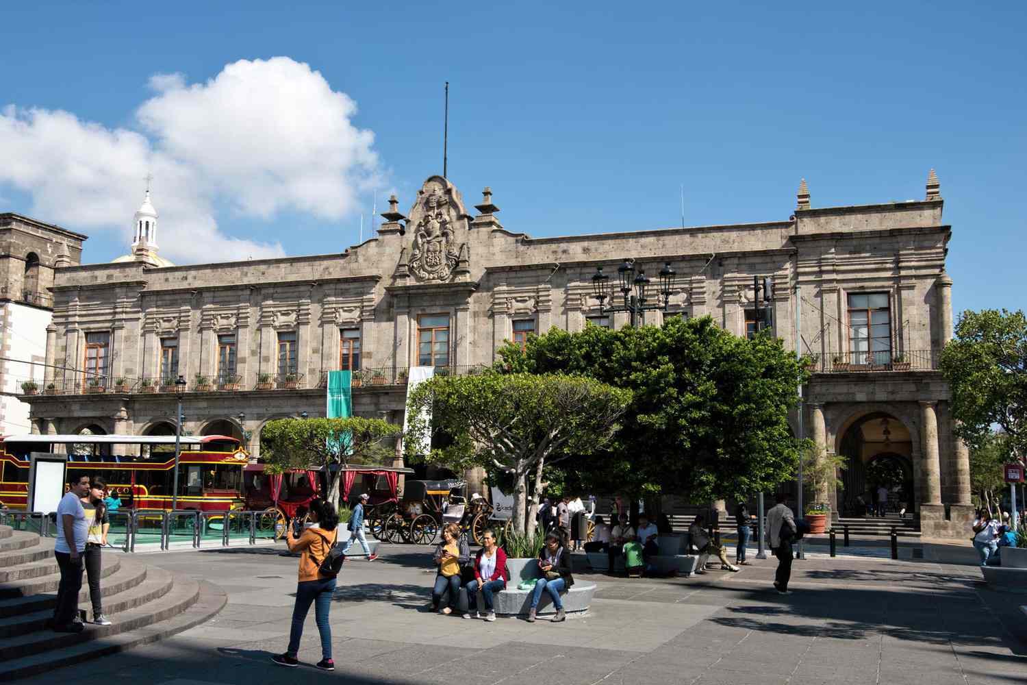 La gente se toma fotos o descansa en los asientos de la Plaza Guadalajara. Al fondo está el Ayuntamiento de Guadalajara (Palacio Municipal de Guadalajara), sede del consejo municipal de la capital del estado de Jalisco, México.