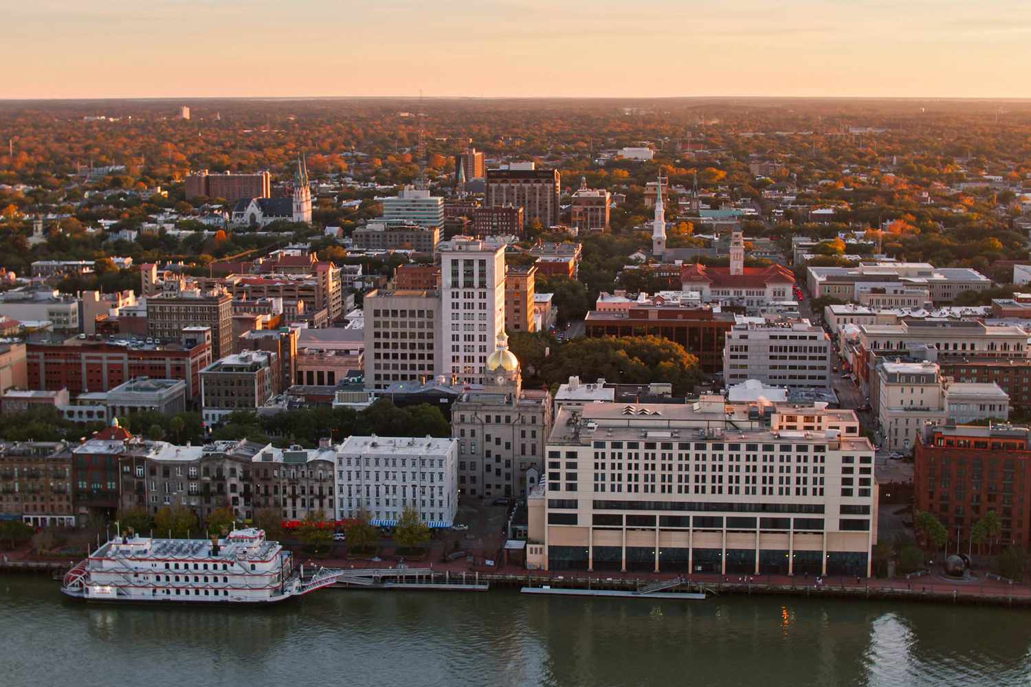 Toma aérea del histórico paseo marítimo de Savannah, Georgia, al atardecer, que incluye un barco fluvial con ruedas de paletas amarrado y la cúpula dorada del ayuntamiento que se eleva sobre los edificios del centro.